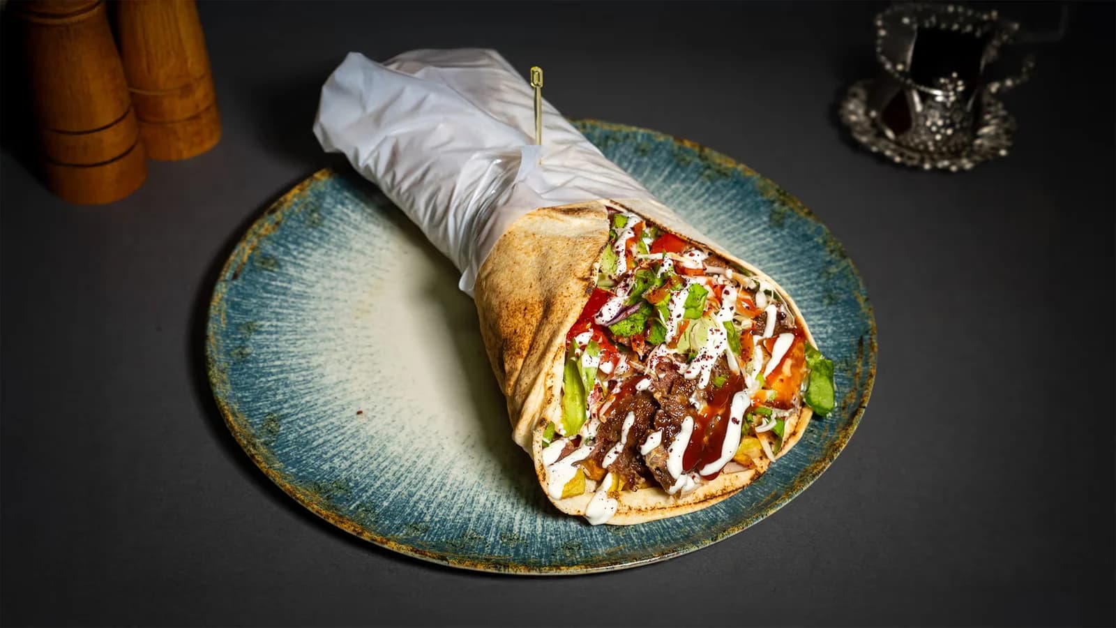 Shawarma in flatbread, wrapped in paper, with meat, vegetables and sauces, on a blue plate